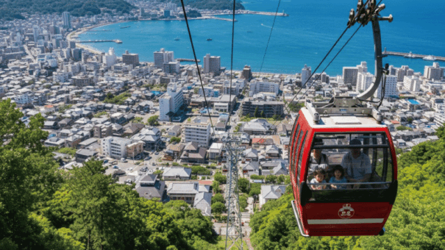 Cheerful daytime photo of the Atami Ropeway gondola going up toward the hillside, with Atami coastline and town visible below. Clear weather, bright natural light, realistic photography style, fun and family-friendly mood.