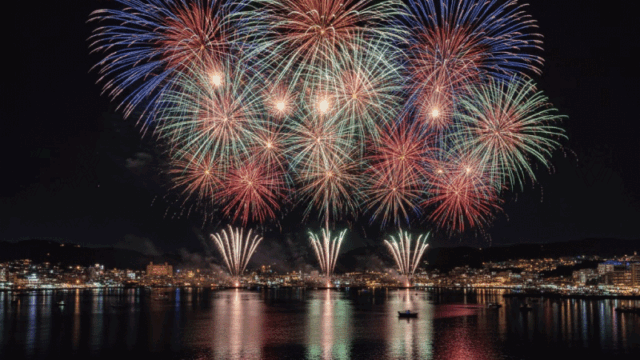 Vibrant yet friendly night photo of fireworks over Atami Bay, reflections on the water, crowds seen only as small silhouettes (no identifiable faces), coastal town lights glowing softly. Realistic photography, festive atmosphere, clean composition, bright and welcoming tone.
