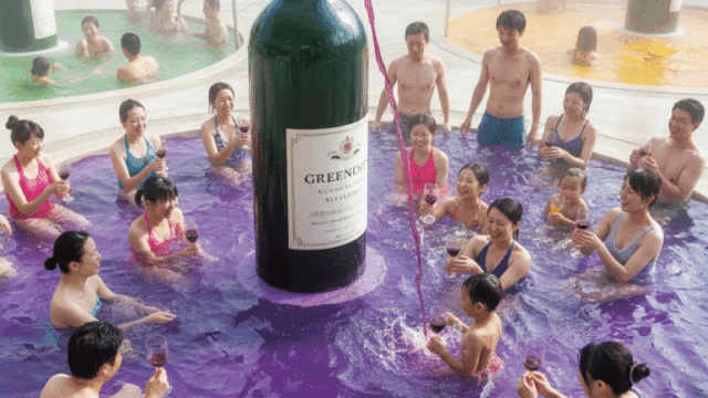 A vibrant photo of a variety of colorful onsens at Hakone Kowakien Yunessun. Shows the famous "Wine Bath" with deep purple water and a giant wine bottle sculpture. People are laughing and enjoying themselves in swimsuits. Fun, bright, and family-friendly atmosphere.