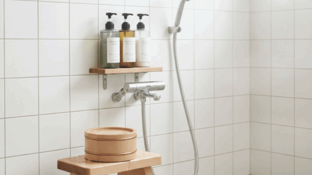 A clean and clear instructional-style photo of an onsen shower station. Shows a small wooden stool, a wooden bucket, and bottled high-end soap/shampoo. Bright white tiles and natural wood, very clean and peaceful atmosphere.