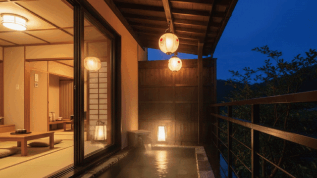 A cozy and romantic high-end photograph of a private outdoor onsen on a wooden balcony at night. Warm glowing lanterns, steam rising into the dark blue evening sky, and a glimpse of a luxury ryokan room with tatami floors. Bright, warm, and high-end travel aesthetic.