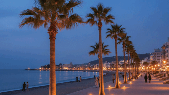 Warm and welcoming evening photo of Atami Sun Beach promenade with palm trees and gentle illuminated lighting, calm sea, people walking in the distance as silhouettes (no close-up faces). Realistic photography, romantic but friendly atmosphere.