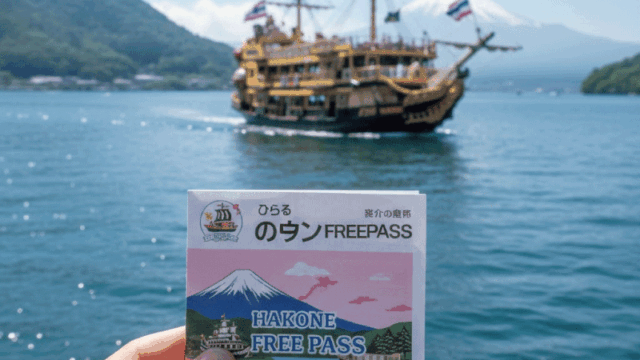 A close-up, first-person perspective of a traveler holding a Hakone Free Pass (digital or paper version) with the iconic Hakone sightseeing "Pirate Ship" on Lake Ashi blurred in the background. Friendly and casual atmosphere, bright sunny day, travel adventure aesthetic.