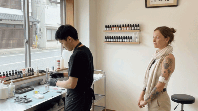 Friendly documentary-style photo of a clean Japanese tattoo studio interior, tattoo artist preparing equipment, foreign customer calmly observing, hygienic and professional atmosphere, soft lighting, modern yet minimal design, realistic photography, welcoming and trustworthy mood